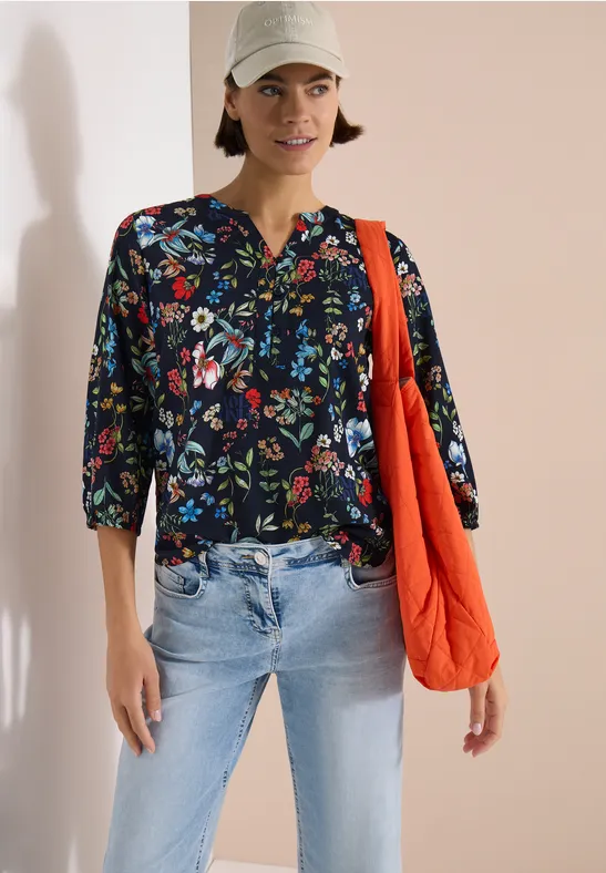 Eine Person trägt eine dunkelblaue Bluse mit Blumenmuster, helle Jeans und eine orangefarbene Tasche.