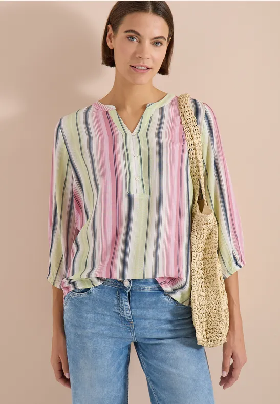 Femme en jean et blouse à fines rayures pastel, col en V, coupe ample, tenant un sac en paille.