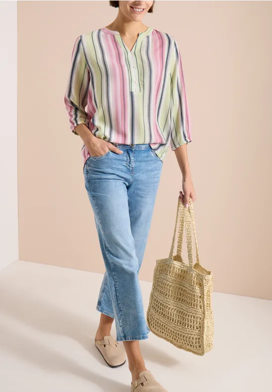 Femme portant un chemisier à rayures pastel, jean bleu et mocassins beiges, tenant un sac en paille tressée.