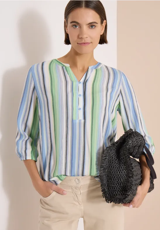 Frau trägt bunte, gestreifte Bluse mit 3/4-Ärmeln, beige Hose und hält eine schwarze Flecht-Tasche.
