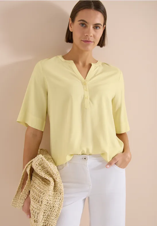 Femme en chemisier jaune pastel à col V et manches mi-longues, pantalon blanc et sac en paille, fond clair