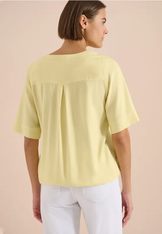 Femme de dos portant un t-shirt jaune pâle à manches courtes et un pantalon blanc.