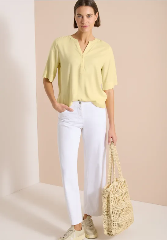 Femme en haut jaune pâle, pantalon blanc, sac en paille et chaussures décontractées sur fond studio épuré.