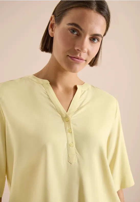Femme en chemisier jaune pastel à col en V et boutons, fond neutre.