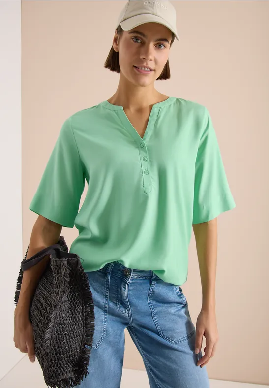 Femme portant un chemisier vert, un jean bleu et tenant un sac tressé noir sur fond beige clair.