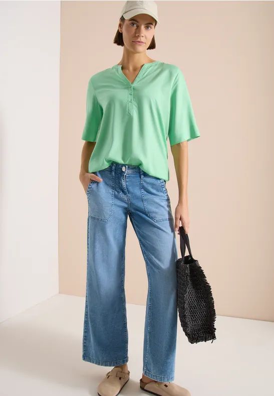 Mannequin portant un chemisier vert pâle à encolure V, un jean large et un sac noir en fausse fourrure sur fond crème.