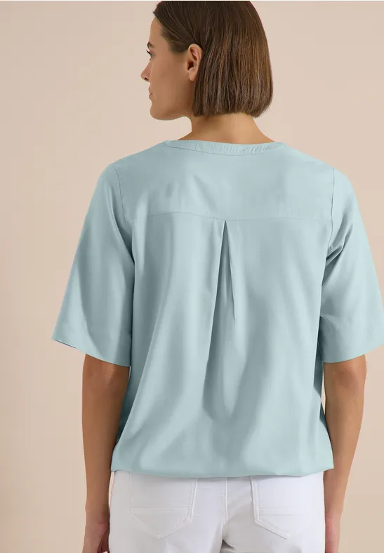 Vue de dos d'une femme aux cheveux mi-longs, portant un chemisier bleu clair à pli au dos et un pantalon blanc.