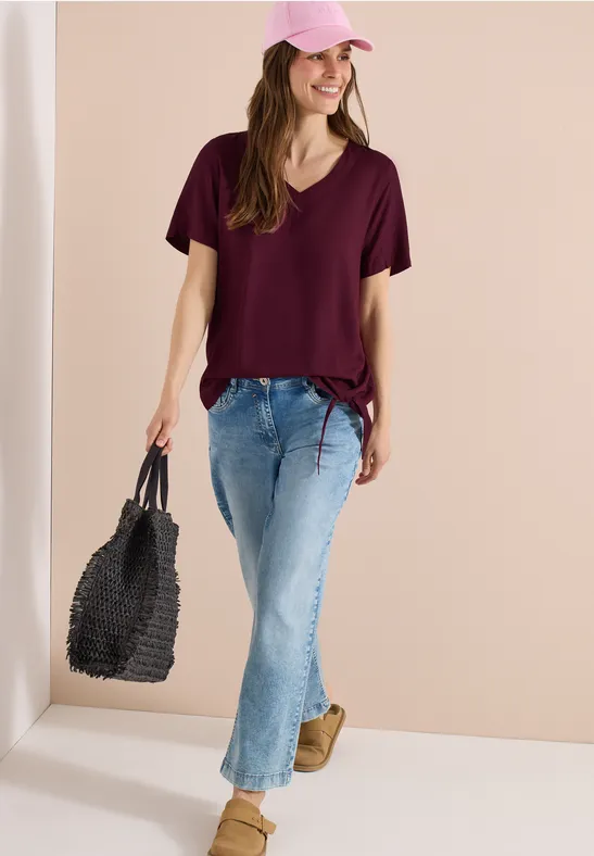 Frau in dunkelrotem T-Shirt, blauen Jeans und beigen Schuhen hält geflochtene schwarze Tasche vor neutralem Hintergrund.