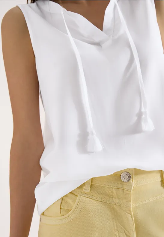 Nahaufnahme einer Person mit ärmellosem weißen Kapuzen-Top mit Kordeln vorne und gelben High-Waist-Hosen.