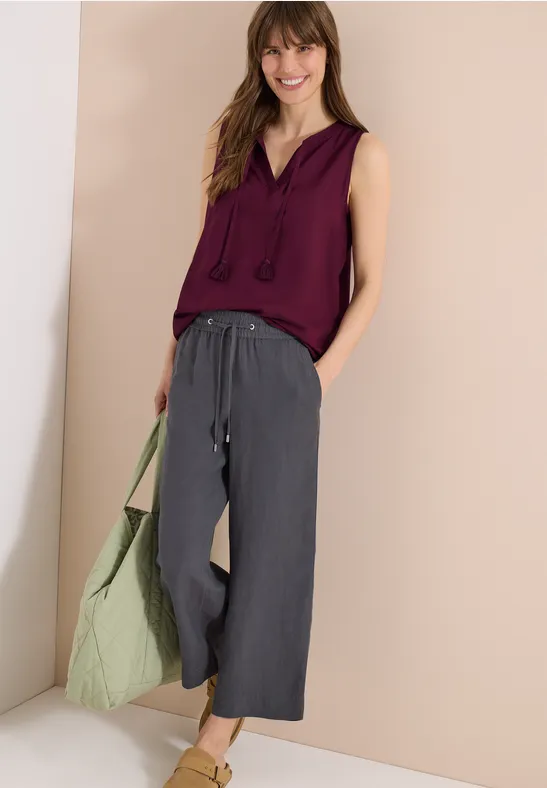 Frau trägt weinrotes ärmelloses Top, weite graue Leinenhose, grüne Tote-Bag und Sandalen.