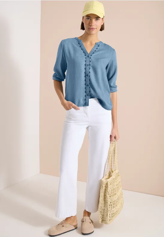 Mannequin porte chemisier bleu clair, pantalon blanc évasé, chaussures beiges et sac en paille.