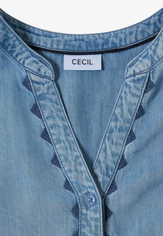 Blouse denim bleu clair à col en V, coutures décoratives triangulaires et étiquette 'CECIL'.