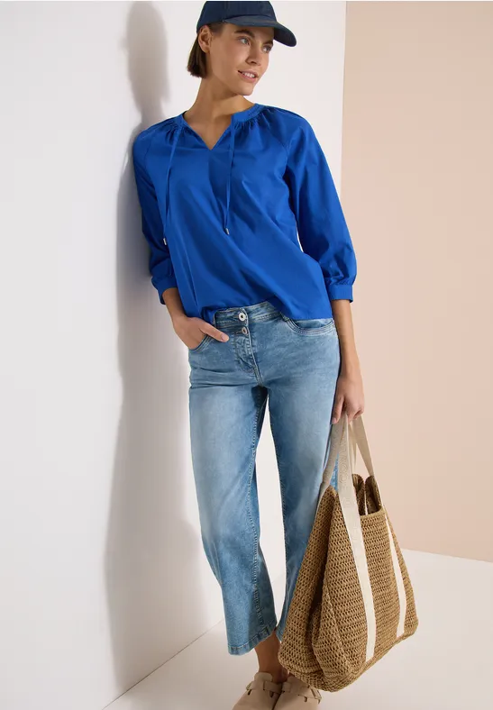 Frau lehnt an Wand mit blauer Bluse, hellen Jeans, Cap und großem geflochtenem Shopper.
