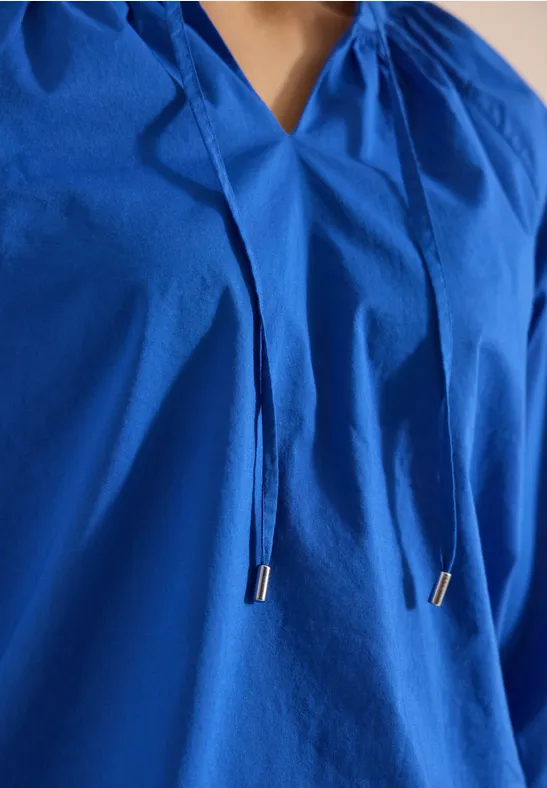 Nahaufnahme einer locker sitzenden, blaufarbenen Bluse mit V-Ausschnitt und Zierbändern am Kragen.