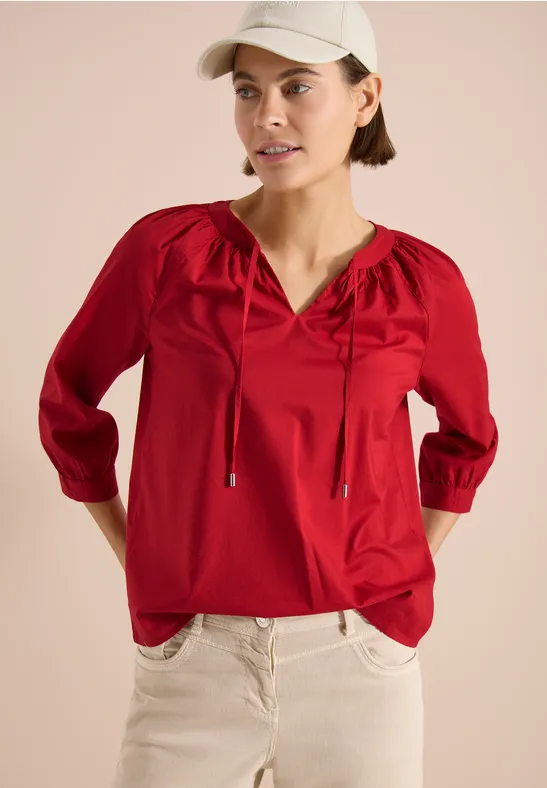 Person in roter Bluse mit 3/4-Ärmeln und Bändchen, kombiniert mit heller Hose und Cap vor beigem Hintergrund.