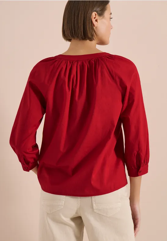 Frau von hinten in roter, locker sitzender Bluse und hellen Hosen vor hellem Hintergrund.