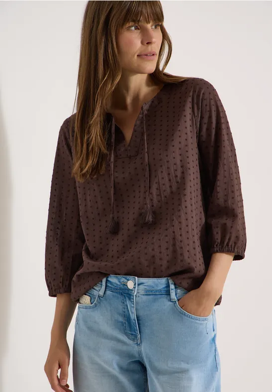 Dame in brauner gepunkteter Bluse mit V-Ausschnitt und Quasten, in hellen Jeans.