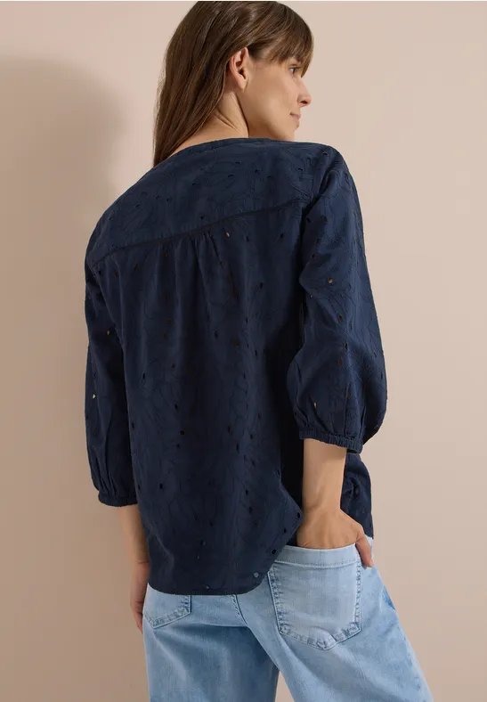 Frau von hinten in dunkelblauer, bestickter Bluse mit Lochstickerei und hellblauen Jeans.