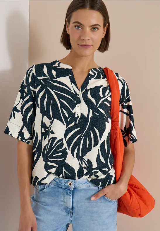 Vrouw met zwart-wit blouseshirt met bladerenprint, lichte jeans en een oranje tas tegen een beige achtergrond.
