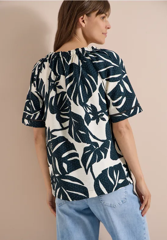 Frau von hinten in weißer Bluse mit dunkelgrünen, großen Tropenblättern und halblangen Ärmeln, dazu hellblaue Jeans.