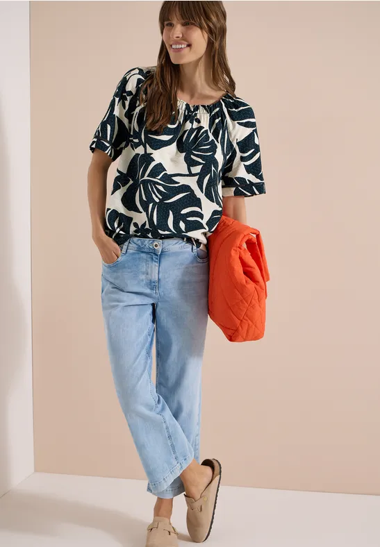 Frau trägt gemustertes Top, helle Jeans und beige Hausschuhe, hält eine orange Jacke in der Hand.