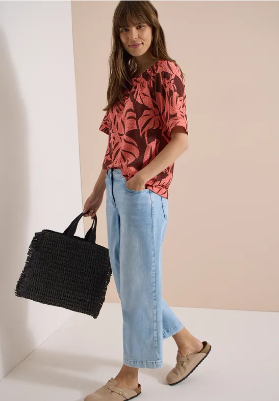 Frau in rotem Tropenprint-Oberteil, hellblauen Culotte-Jeans, beigen Slip-On-Schuhen und schwarzer Korbtasche.