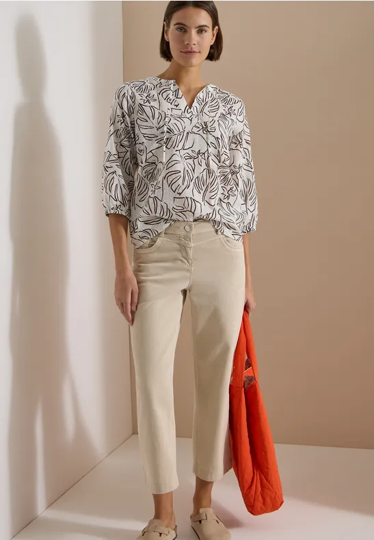 Personne en chemise blanche à motifs, pantalon clair et grand sac orange, posant devant un mur beige.