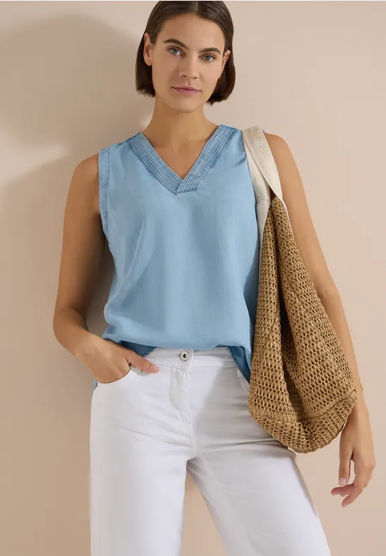Femme portant un top bleu clair sans manches col V, pantalon blanc et grand sac en paille sur fond neutre.