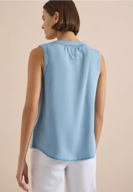 Femme de dos portant un top denim sans manches bleu clair et un pantalon blanc.