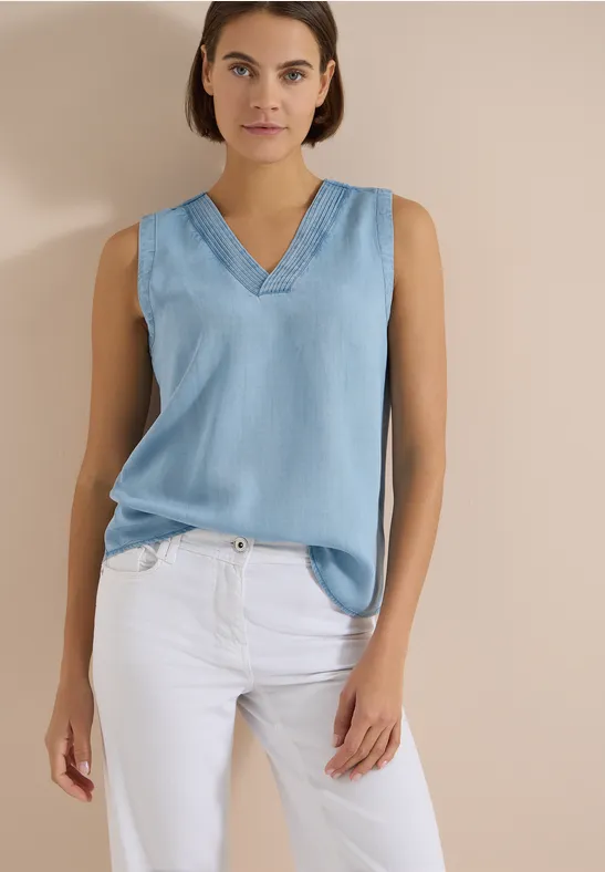 Femme devant un fond beige, portant un top sans manches bleu clair à col V et un pantalon blanc.