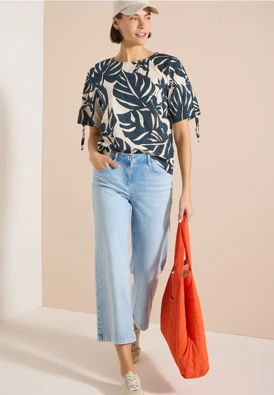 Frau trägt eine Bluse mit Blättermuster, helle Schlagjeans, grüne Sneaker und rote Tasche.