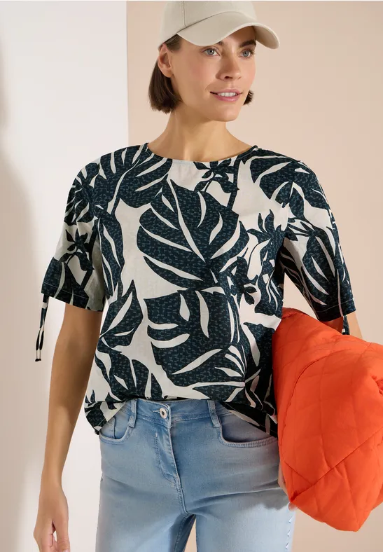 Frau in weißem T-Shirt mit blauem Blattmuster, hellen Jeans und oranger Tasche.
