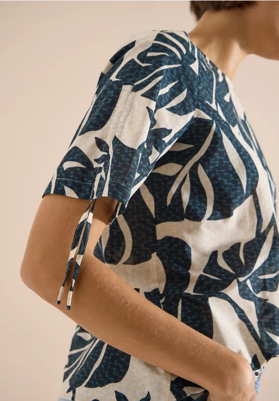 Nahaufnahme einer Frau in weiß-blauem Blatt-Print-Shirt mit kurzen Ärmeln und taillierter, heller Hose.