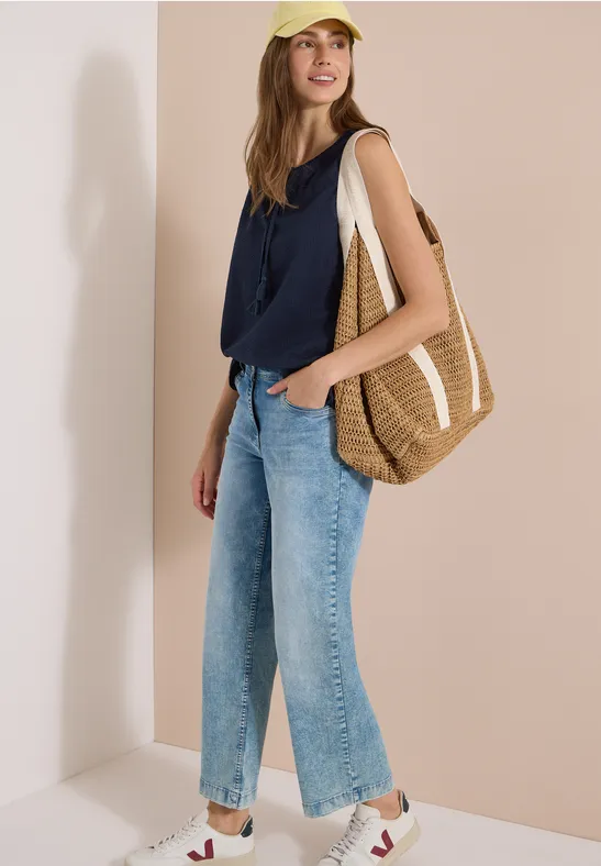 Femme appuyée contre un mur beige, portant un débardeur bleu marine, jean large, baskets blanches et grand sac tressé.
