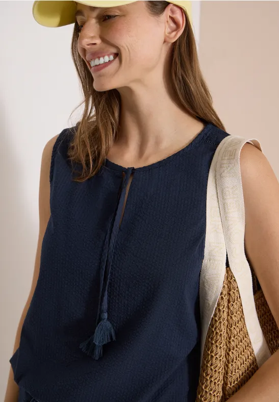 Femme en top bleu marine sans manches à pompons, portant deux sacs d’épaule en toile et paille.