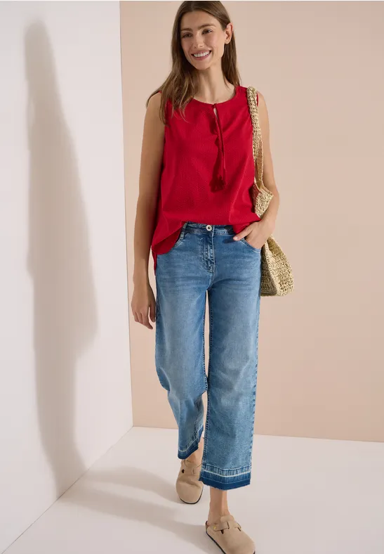 Femme en top rouge sans manches, jean bleu ample et sac tressé en paille devant un mur clair.