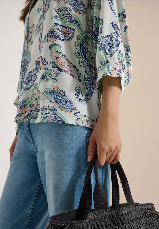 Personne en chemisier à motif cachemire et jean tient un sac noir tressé.