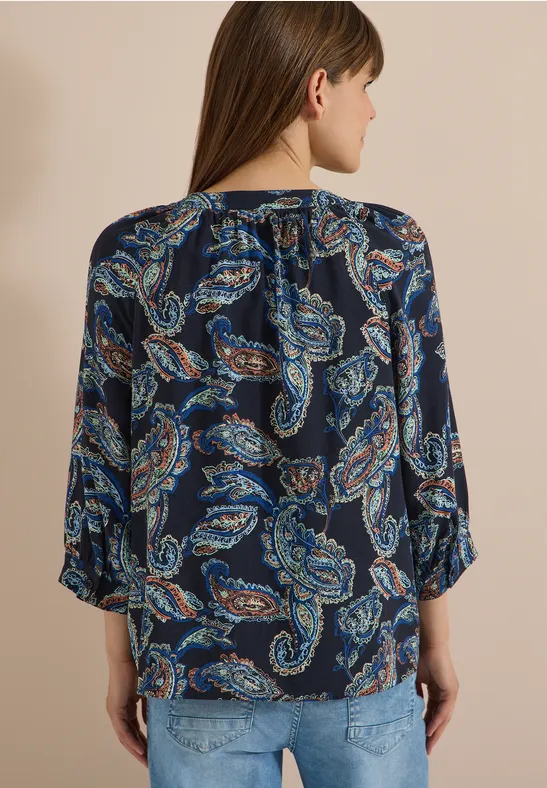 Frau von hinten in dunkelblauer Bluse mit Paisley-Print und Jeans vor beigem Hintergrund.
