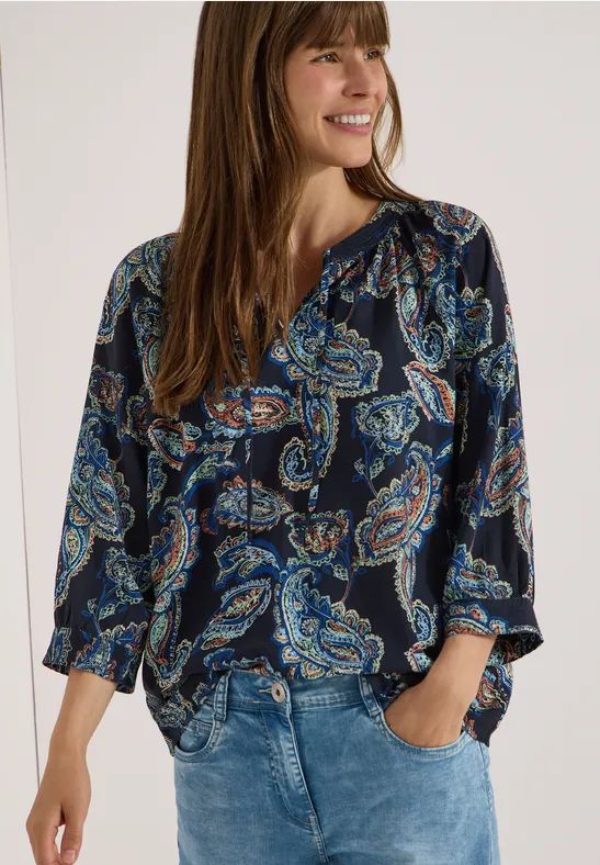 Frau in Bluse mit Paisley-Print und Jeans, Hände in den Taschen.