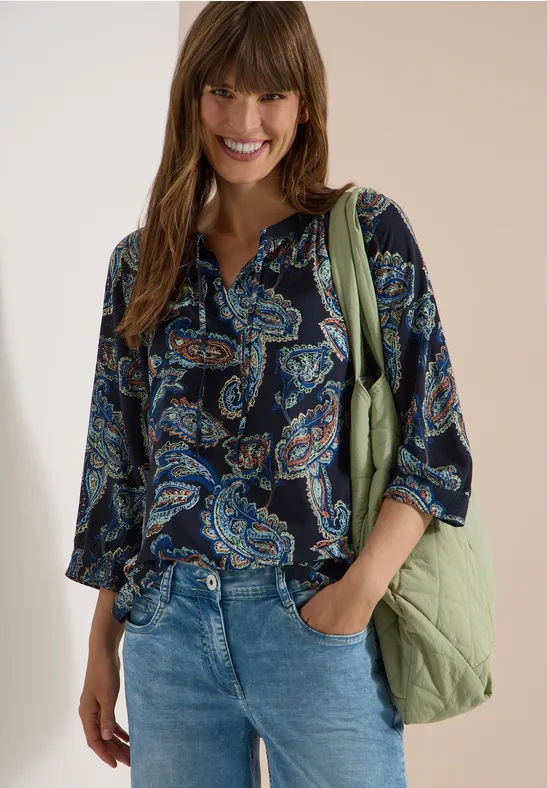 Frau in dunkler Paisley-Bluse, Jeans und grüner Tasche, entspannt stehend.