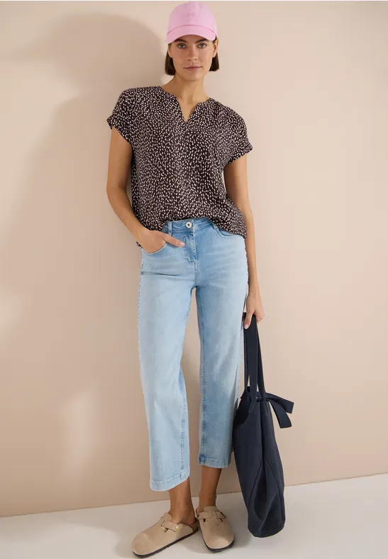 Frau trägt gemustertes Kurzarm-Shirt, hellblaue Straight-Jeans, bequeme Slipper und hält eine dunkelblaue Tragetasche.