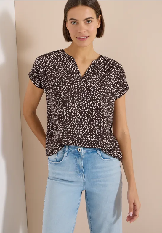 Frau in gemusterter Bluse und heller Jeans steht mit Händen in den Taschen vor hellem Hintergrund.