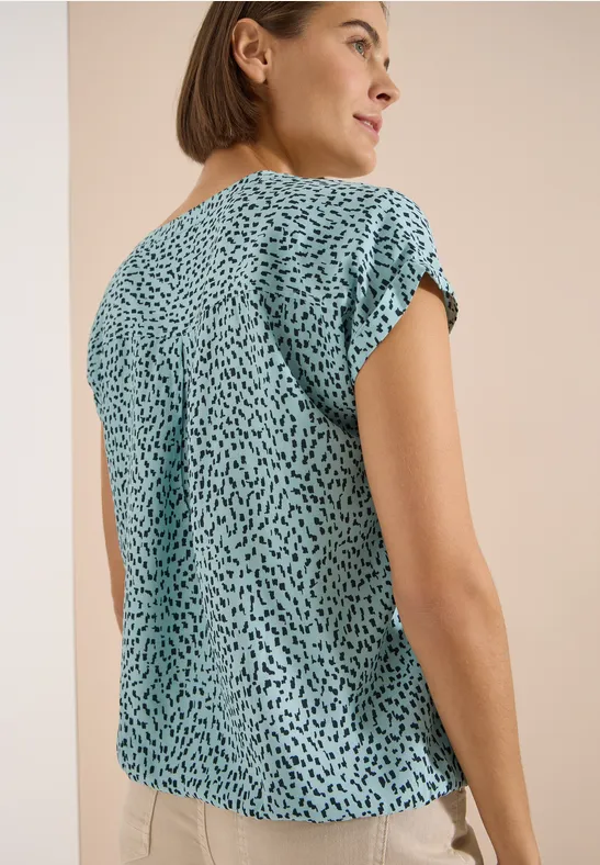 Femme de dos portant un t-shirt turquoise à manches courtes avec un motif à gouttes noires.