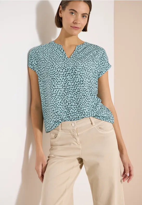 Une femme porte un chemisier à motifs verts et un pantalon beige devant un mur clair.