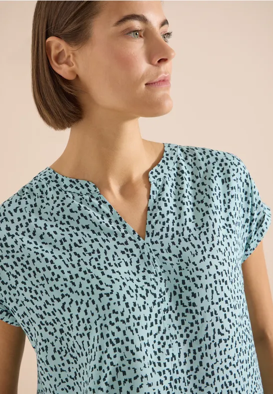 Femme portant un haut turquoise à motifs noirs abstraits, encolure en V et manches courtes.