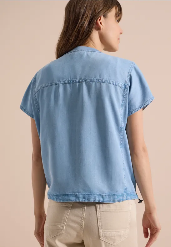 Frau von hinten in hellblauer, kurzärmeliger Jeansbluse und hellen Hosen.