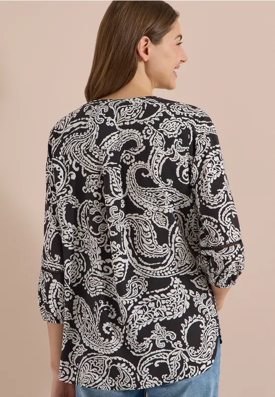 Femme de dos portant un haut noir à motif cachemire blanc sur fond beige.