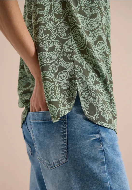 Seitliche Ansicht einer Person mit grün-weiß paisleygemustertem Hemd und blauen Jeans, Hand in der Tasche.