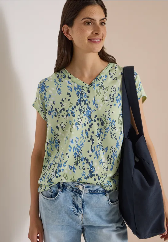 Femme souriante en chemisier vert à fleurs et jean, avec un sac bleu porté sur l'épaule.