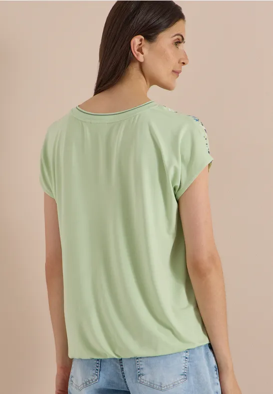 Femme vue de dos portant un top vert clair à manches courtes avec motif aux épaules et jean bleu.